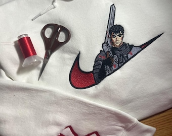 May include: White hoodie with a red and black embroidered design featuring a character with a sword. The design is incorporated into a swoosh logo. A red thread spool and scissors are also visible.