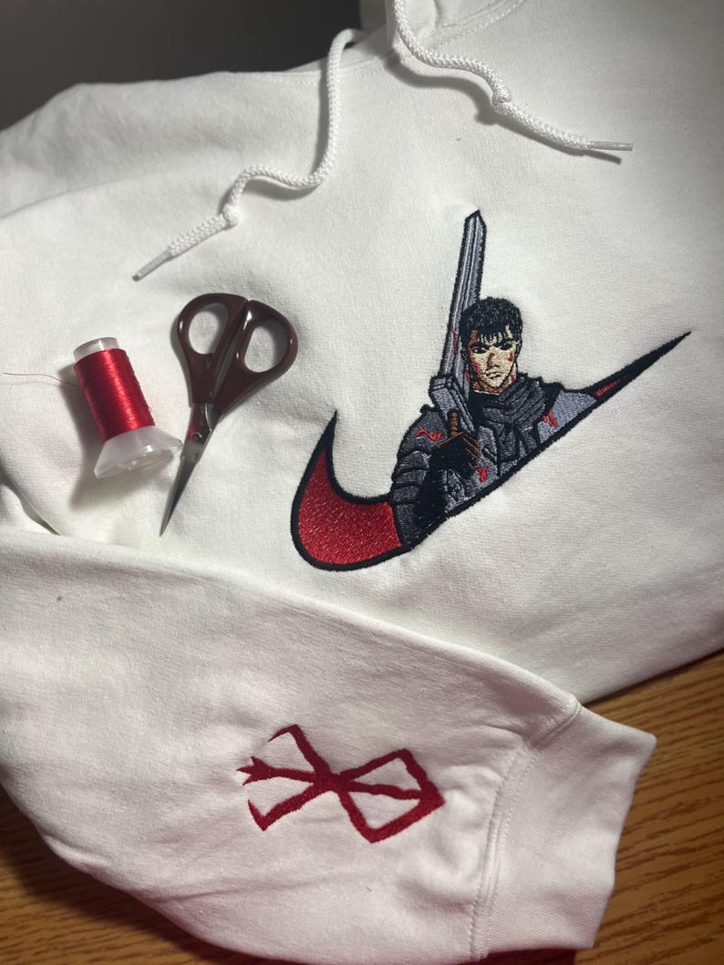 May include: White hoodie with a red and black embroidered design featuring a character with a sword. The design is incorporated into a swoosh logo. A red thread spool and scissors are also visible.