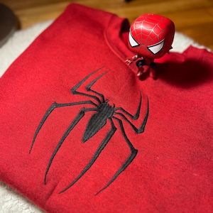Może przedstawiać: Czerwona bluza z dużym, czarnym emblematem pająka z przodu. Figurka Spider-Mana umieszczona na ramieniu. Bluza jest złożona na białej, teksturowanej powierzchni.