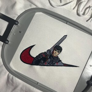 May include: White hoodie with an embroidered design. The design features a figure holding a sword, integrated into a red and black swoosh. The embroidery is detailed, with a focus on the character's armor and expression. The hoodie's drawstring is visible.