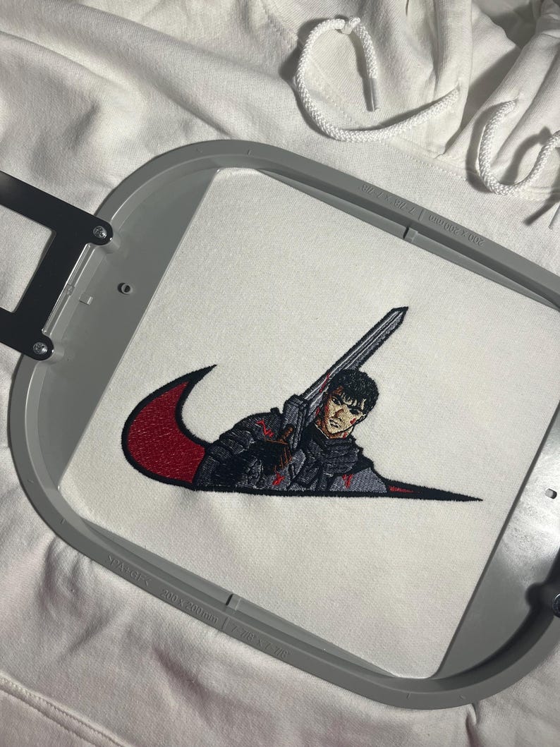 May include: White hoodie with an embroidered design. The design features a figure holding a sword, integrated into a red and black swoosh. The embroidery is detailed, with a focus on the character's armor and expression. The hoodie's drawstring is visible.