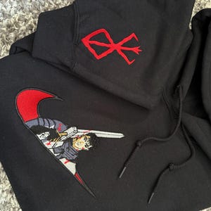May include: Black hoodie with a red embroidered design on the upper left chest and a large embroidered graphic on the front. The front graphic features a character with a sword. The hoodie has a drawstring hood.