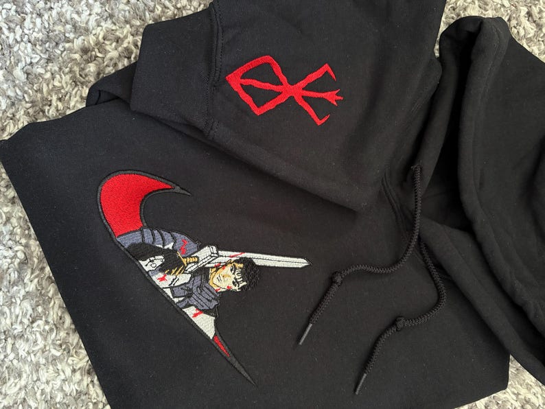 May include: Black hoodie with a red embroidered design on the upper left chest and a large embroidered graphic on the front. The front graphic features a character with a sword. The hoodie has a drawstring hood.