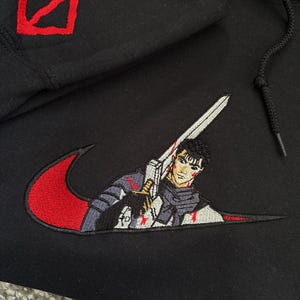 May include: Black hoodie with red and white embroidered design. Features a red symbol on the hood and a stylized image of a character holding a sword within a red swoosh.
