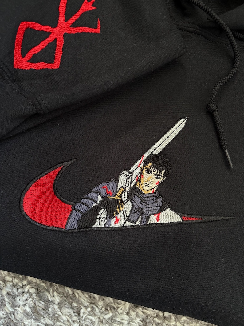 May include: Black hoodie with red and white embroidered design. Features a red symbol on the hood and a stylized image of a character holding a sword within a red swoosh.