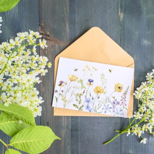 May include: A white card with a watercolor illustration of yellow and blue wildflowers and bees. The card is in a brown envelope and surrounded by white flowers.