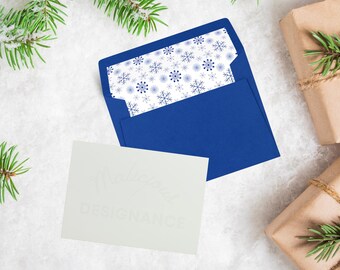 May include: A royal blue envelope with a snowflake-patterned liner and a white card with the text "Malicious Designance" in a soft, light font. The scene includes wrapped gifts and evergreen branches on a snowy surface.