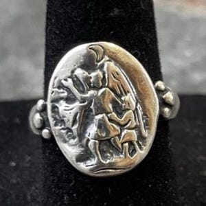 Puede incluir: Anillo plateado con un centro ovalado que presenta un relieve detallado de figuras. El anillo tiene un dise&ntilde;o decorativo en los laterales y se muestra sobre un soporte negro. El dise&ntilde;o del anillo sugiere un tema hist&oacute;rico o religioso.