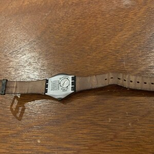 Op de afbeelding: Een Swatch-horloge met een bruine leren band en een zwarte gesp. De wijzerplaat van het horloge is zilverkleurig met zwarte tekst en een zilveren wijzerplaat. Het horloge ligt op een houten oppervlak.