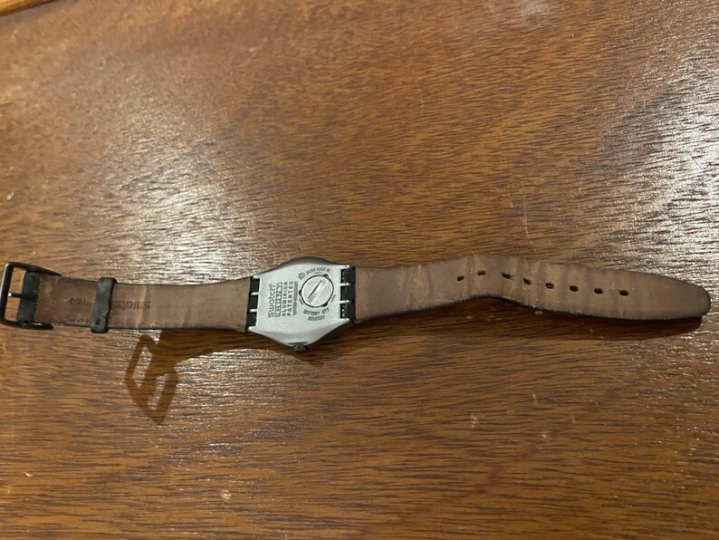 Op de afbeelding: Een Swatch-horloge met een bruine leren band en een zwarte gesp. De wijzerplaat van het horloge is zilverkleurig met zwarte tekst en een zilveren wijzerplaat. Het horloge ligt op een houten oppervlak.