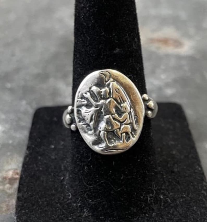 Puede incluir: Anillo plateado con un centro ovalado que presenta un relieve detallado de figuras. El anillo tiene un dise&ntilde;o decorativo en los laterales y se muestra sobre un soporte negro. El dise&ntilde;o del anillo sugiere un tema hist&oacute;rico o religioso.