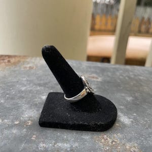 Puede incluir: Un anillo de plata con un dise&ntilde;o detallado, exhibido en un soporte negro para anillos. El intrincado patr&oacute;n del anillo es visible, mostrando su artesan&iacute;a. El anillo se encuentra sobre un fondo borroso al aire libre.