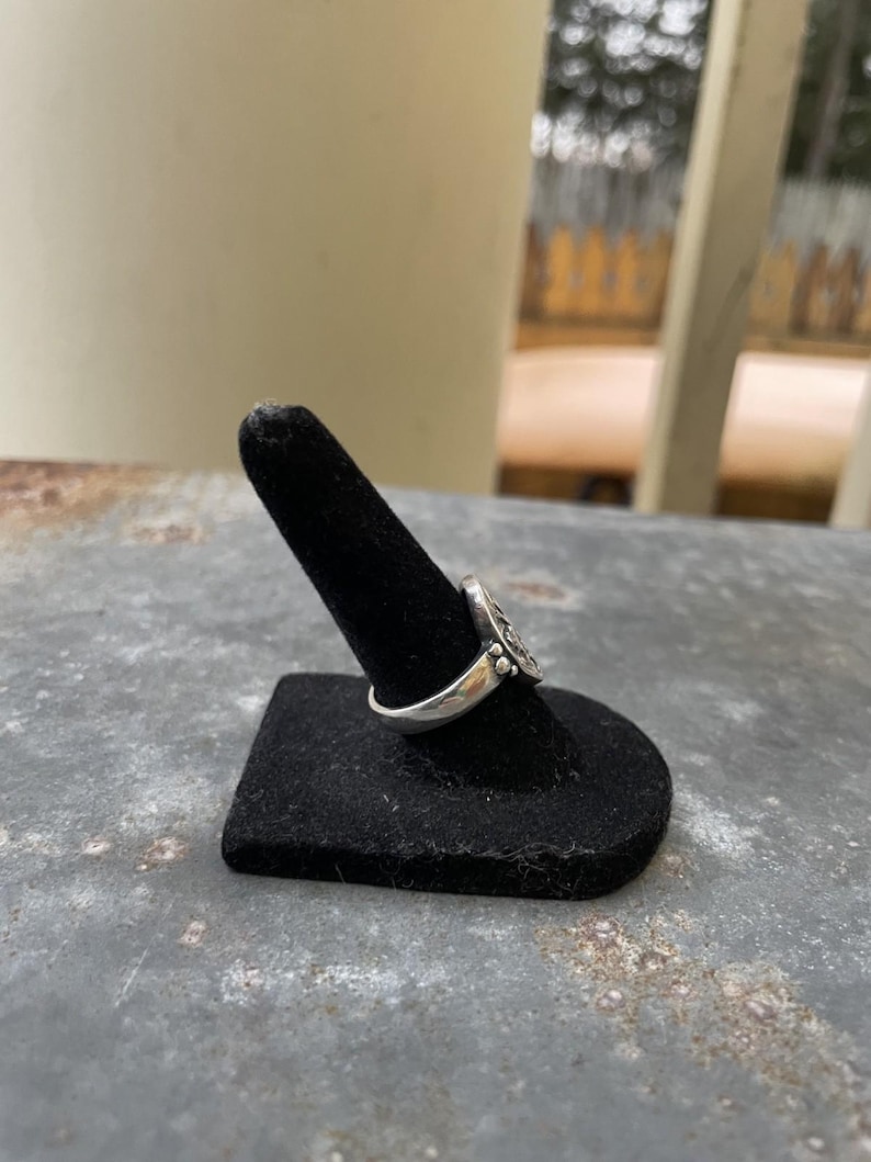 Puede incluir: Un anillo de plata con un dise&ntilde;o detallado, exhibido en un soporte negro para anillos. El intrincado patr&oacute;n del anillo es visible, mostrando su artesan&iacute;a. El anillo se encuentra sobre un fondo borroso al aire libre.