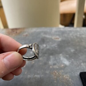 Puede incluir: Un anillo de plata con una cara ovalada detallada con un dise&ntilde;o en relieve. El anillo est&aacute; sujeto por los dedos de una persona. La cara del anillo tiene un patr&oacute;n detallado y el metal tiene un aspecto ligeramente envejecido.