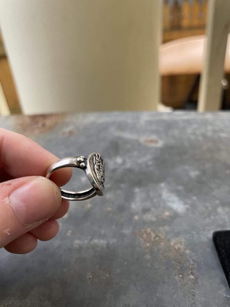 Puede incluir: Un anillo de plata con una cara ovalada detallada con un dise&ntilde;o en relieve. El anillo est&aacute; sujeto por los dedos de una persona. La cara del anillo tiene un patr&oacute;n detallado y el metal tiene un aspecto ligeramente envejecido.
