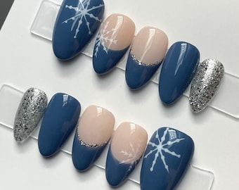 Puede incluir: Un juego de diez uñas artificiales con varios diseños. Algunas uñas son de color azul marino liso, otras tienen una base nude con un acento de purpurina plateada o un diseño de copo de nieve blanco. Las uñas tienen forma ovalada.
