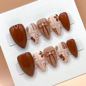 May include: A set of almond-shaped press-on nails in various shades of brown. Some nails feature a solid, glossy finish, while others showcase designs including bows, flowers, and bear illustrations. The nails are arranged on a clear plastic display.
