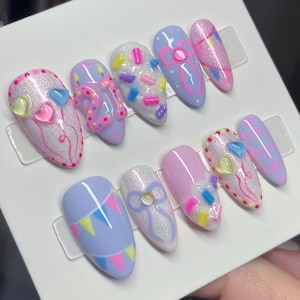 May include: A set of ten almond-shaped press-on nails featuring a pastel color palette of lavender, pink, and white. Designs include raised 3D sprinkles, bows, heart-shaped charms, birthday candles, the number 21, and colorful bunting flags.