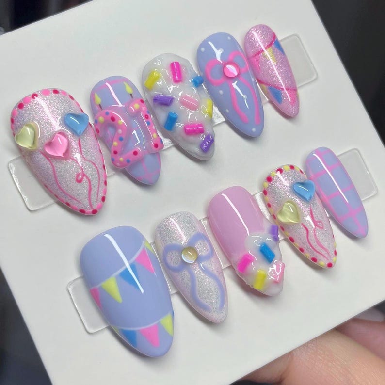 May include: A set of ten almond-shaped press-on nails featuring a pastel color palette of lavender, pink, and white. Designs include raised 3D sprinkles, bows, heart-shaped charms, birthday candles, the number 21, and colorful bunting flags.