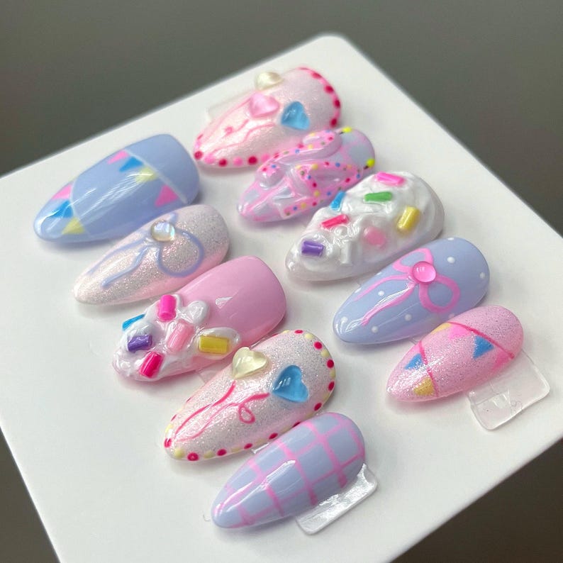 May include: A set of ten almond-shaped press-on nails featuring pastel colors, glitter finishes, and 3D embellishments. Designs include pink and blue bows, white icing textures with colorful sprinkles, heart accents, and geometric patterns.