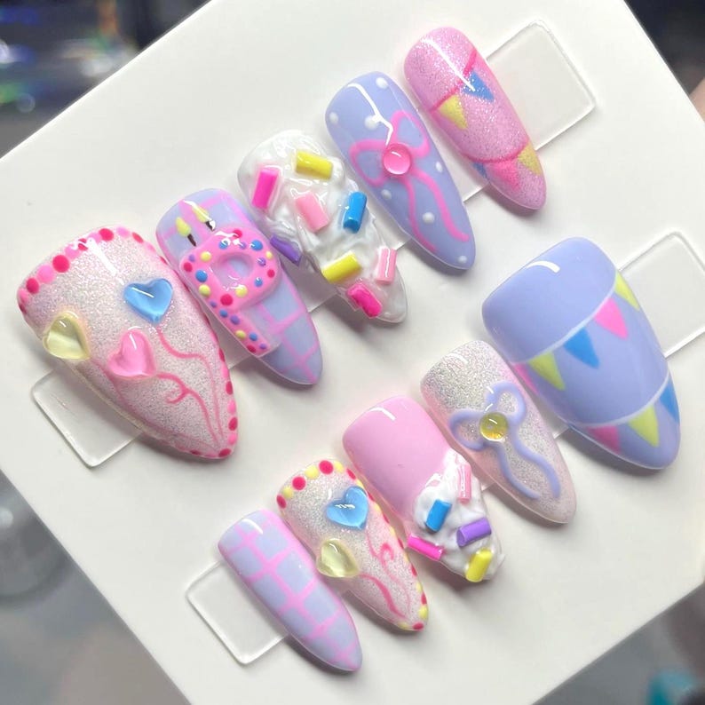 May include: A set of ten almond-shaped press-on nails featuring a pastel color palette of pink, lavender, and white. The designs include raised 3D elements like colorful sprinkles, bows, heart-shaped balloons, and festive bunting patterns.