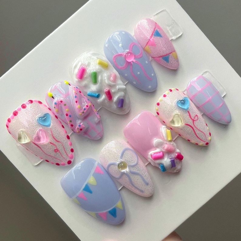 May include: A set of ten press-on nails featuring a pastel color palette of pink, blue, and white. Designs include raised 3D sprinkles, hand-painted bows, colorful bunting flags, grid patterns, and small translucent heart-shaped embellishments.