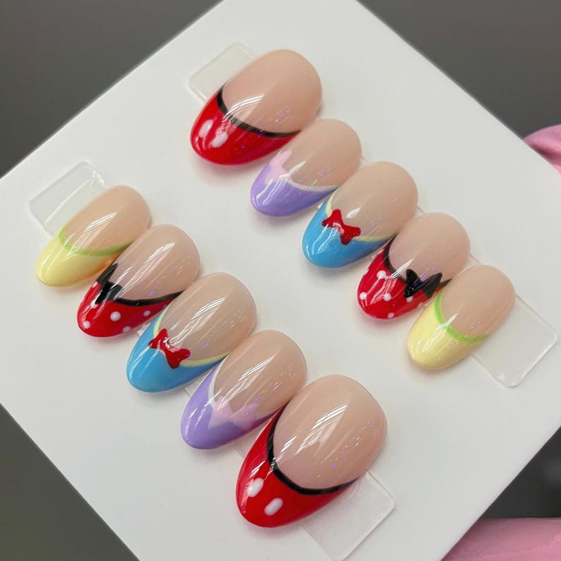 May include: Set of ten false nails with different Disney character designs. The nails are oval shaped and have a light pink base with a white tip. The designs include a red nail with white polka dots and a black bow, a blue nail with a yellow stripe and a red bow, a purple nail with a white stripe and a red bow, a yellow nail with a green stripe and a black bow, and a red nail with a black stripe and white polka dots.