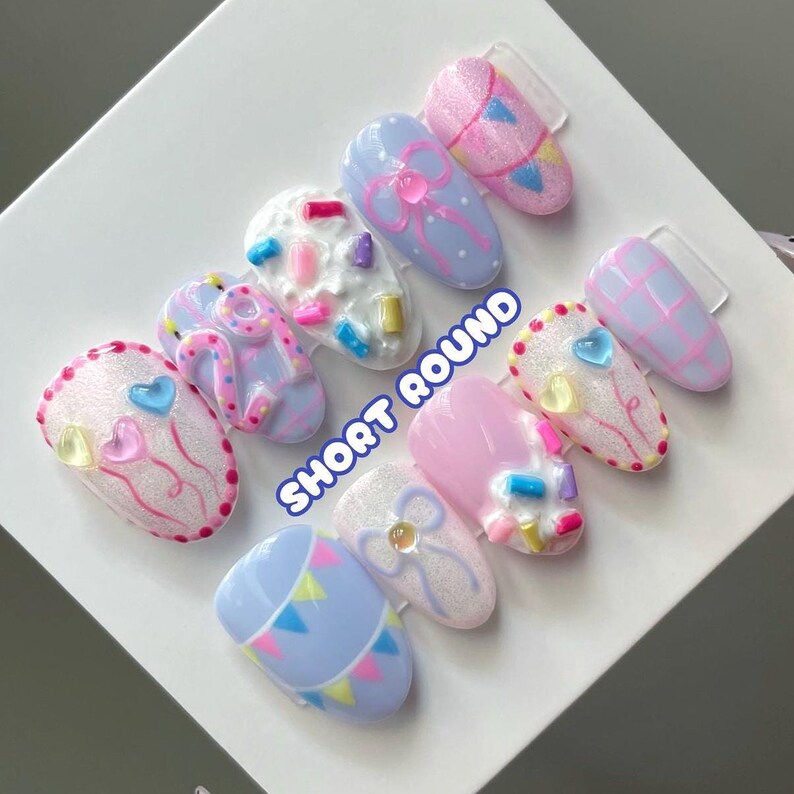 May include: A set of ten short round press-on nails featuring pastel colors, glitter textures, and 3D embellishments including bows, hearts, and sprinkles. The designs include polka dots, plaid patterns, and bunting flags with the text SHORT ROUND.