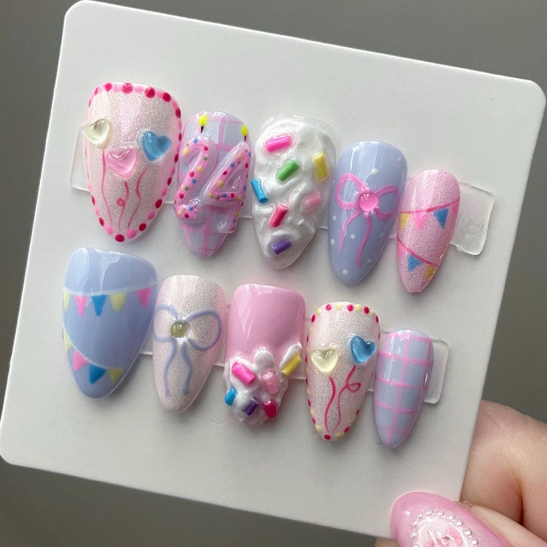 May include: A set of ten almond-shaped press-on nails featuring a birthday theme. Designs include pink and blue plaid, white frosting with colorful sprinkles, light blue with bunting, pink with bows, and heart balloons with the number 24.