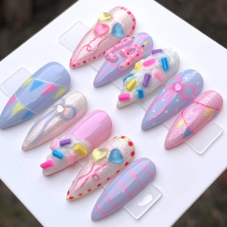 May include: A set of ten almond-shaped press-on nails featuring a pastel color palette of light blue, pink, and white. Designs include hand-painted bunting, glitter finishes, raised 3D sprinkles, heart-shaped charms, and plaid patterns.