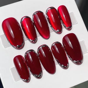 May include: Set of ten oval-shaped, glossy, deep red press-on nails with a silver metallic trim. The nails are arranged on a white background, showcasing their vibrant color and design. A stylish manicure accessory.