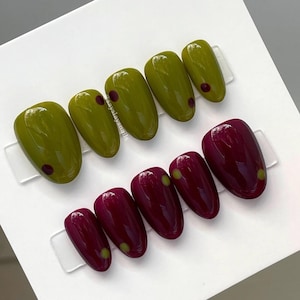 May include: A set of ten press-on nails with a glossy finish. The top row features four olive green nails with small dark red dots. The bottom row features six deep burgundy nails with small olive green dots. The text says sayslaysnails.