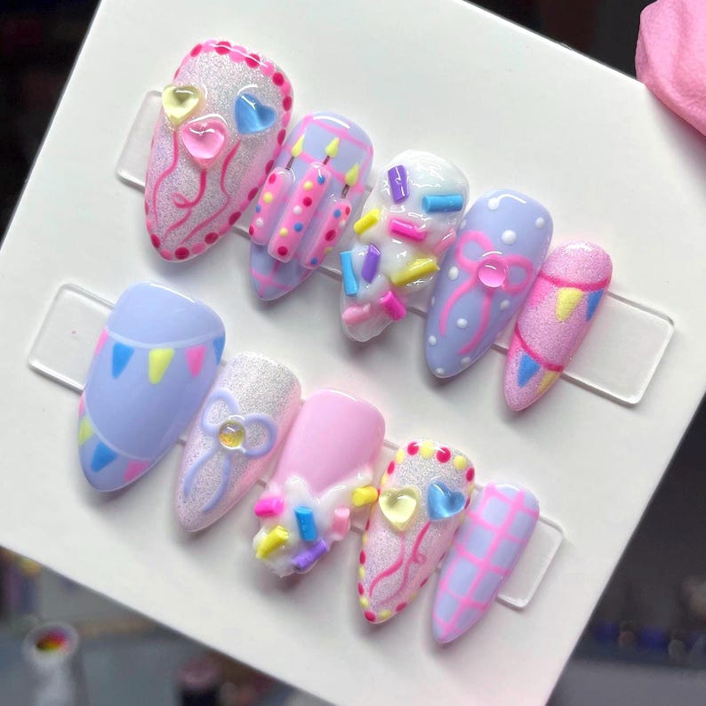 May include: A set of ten press-on nails featuring a birthday theme with pastel colors. Designs include pink and blue heart charms, raised birthday candles, colorful sprinkles, bows with center gems, bunting flags, and a pink grid pattern on a white base.