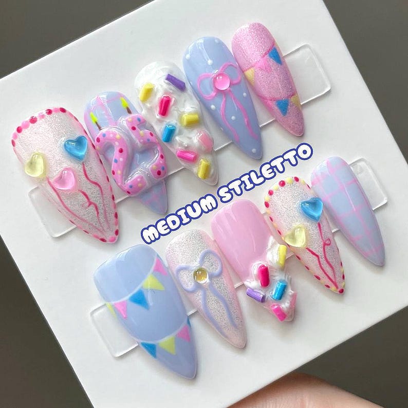 May include: A set of ten medium stiletto press-on nails featuring a pastel color palette of light blue, pink, and white. Designs include 3D number 25, colorful sprinkles, bows, bunting flags, hearts, and plaid patterns with a glittery finish.
