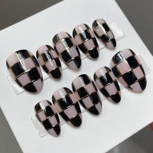 May include: Set of ten almond-shaped false nails. Each nail features a checkerboard pattern in black and light pink, outlined with gold metallic lines. The nails are arranged on clear plastic strips against a white background.