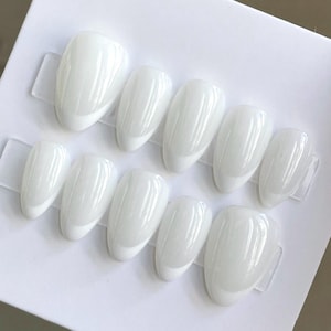 May include: A set of ten almond-shaped press-on nails with a glossy, solid white finish. The nails are arranged in two rows of five on a white display card, featuring a smooth, reflective surface and a rounded, tapered tip design.