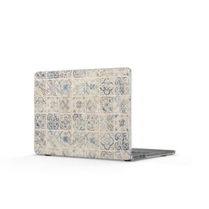 May include: A laptop with a protective skin featuring a repeating grid pattern of square tiles. The tiles display intricate floral and geometric motifs in muted shades of blue, beige, and cream, creating a weathered, aged appearance across the surface.