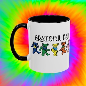 May include: White ceramic coffee mug with a black rim and handle. The mug features a graphic of the Grateful Dead dancing bears in rainbow colors and the text "Grateful Dead".