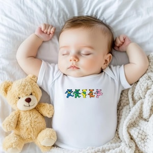 May include: A sleeping baby wearing a white onesie with a colorful bear graphic. A tan teddy bear is next to the baby on a white sheet. The bear graphic features five dancing bears in blue, green, yellow, orange, and pink.