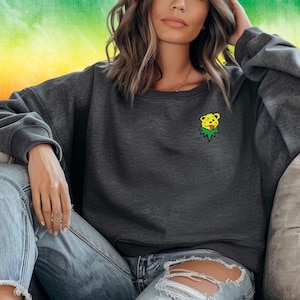 May include: A dark gray sweatshirt with a yellow bear face graphic on the left chest. The bear is wearing a green leafy crown and has a tongue sticking out. The sweatshirt is worn by a person sitting on a couch.
