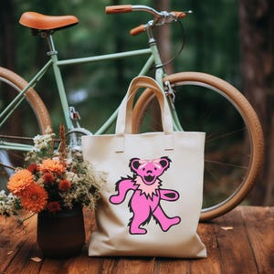 May include: Cream-colored tote bag featuring a pink dancing bear graphic with a pink bow and a ruffled collar. A green bicycle with a brown seat and tires is in the background. A vase of orange and white flowers sits on a wooden surface.