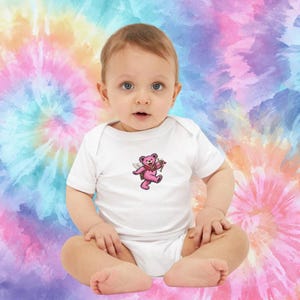 May include: A white baby onesie with a pink bear graphic wearing a bow tie and wings. The baby is sitting in front of a colorful tie-dye background. The onesie is short-sleeved and has a simple, classic design.