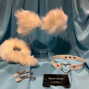 Può includere: Fascia per capelli con orecchie di gatto in pelliccia sintetica bianca, coda e collare con ciondolo a forma di cuore argentato. Il collare è in similpelle bianca e presenta dettagli in metallo argentato. Il set include anche due morsetti in metallo argentato. Il testo sulla carta recita "Discreet London Est. 2022 DiscreetLondon.uk"