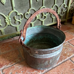 May include: A small, antique copper bucket with a rusty metal handle. The bucket has a round shape with a slightly flared rim and a patinaed finish, showing shades of brown and green. The handle is arched and attached to the sides.
