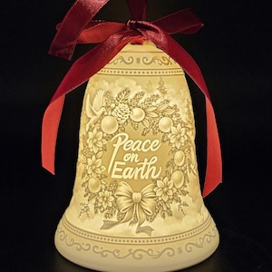 May include: Cream-colored ceramic bell with a red satin ribbon. The bell features a floral and fruit design with the words "Peace on Earth" in the center. A decorative bow is at the bottom of the bell.