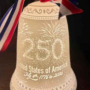 May include: A white porcelain bell ornament featuring a red, white, and blue ribbon. The bell is etched with the number 250, the text United States of America, and July 4th, 1776 to 2026, surrounded by firework and star patterns.