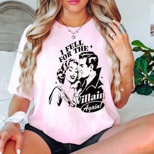 May include: Pink t-shirt with a black and white illustration of a couple. The text on the shirt reads "I Fell For The Villain Again!"