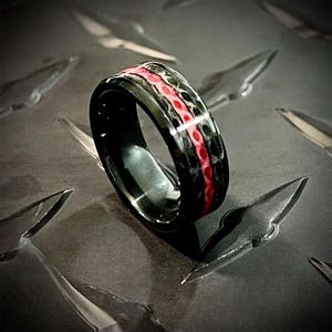 May include: A black ring with a red inlay. The ring has a hammered texture.