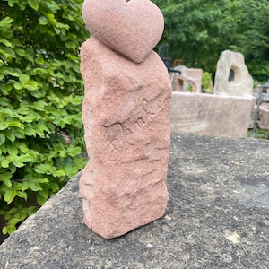 Könnte beinhalten: Eine rotbraune Steinskulptur, die ein Herz auf einem strukturierten, rechteckigen Sockel zeigt. Das Wort "Danke" ist in den Sockel gemeißelt. Die Skulptur befindet sich im Freien, vor dem Hintergrund von grünem Laub und anderen Steinstrukturen.