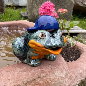 Könnte beinhalten: Eine Keramikfrosch-Skulptur mit einer blauen Baseballkappe. Der Frosch hat eine grün-schwarze, gesprenkelte Haut, einen orangefarbenen Mund und Füße sowie große, ausdrucksstarke Augen. Er sitzt auf einem rotbraunen Stein, in der Nähe von Wasser und rosa Blüten.